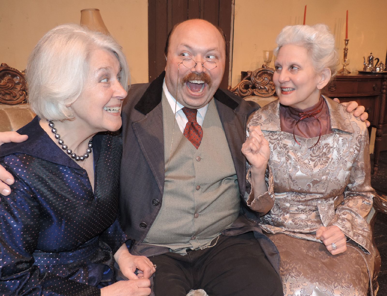 Two white-haired women in dresses sit with a bald man in a suit.