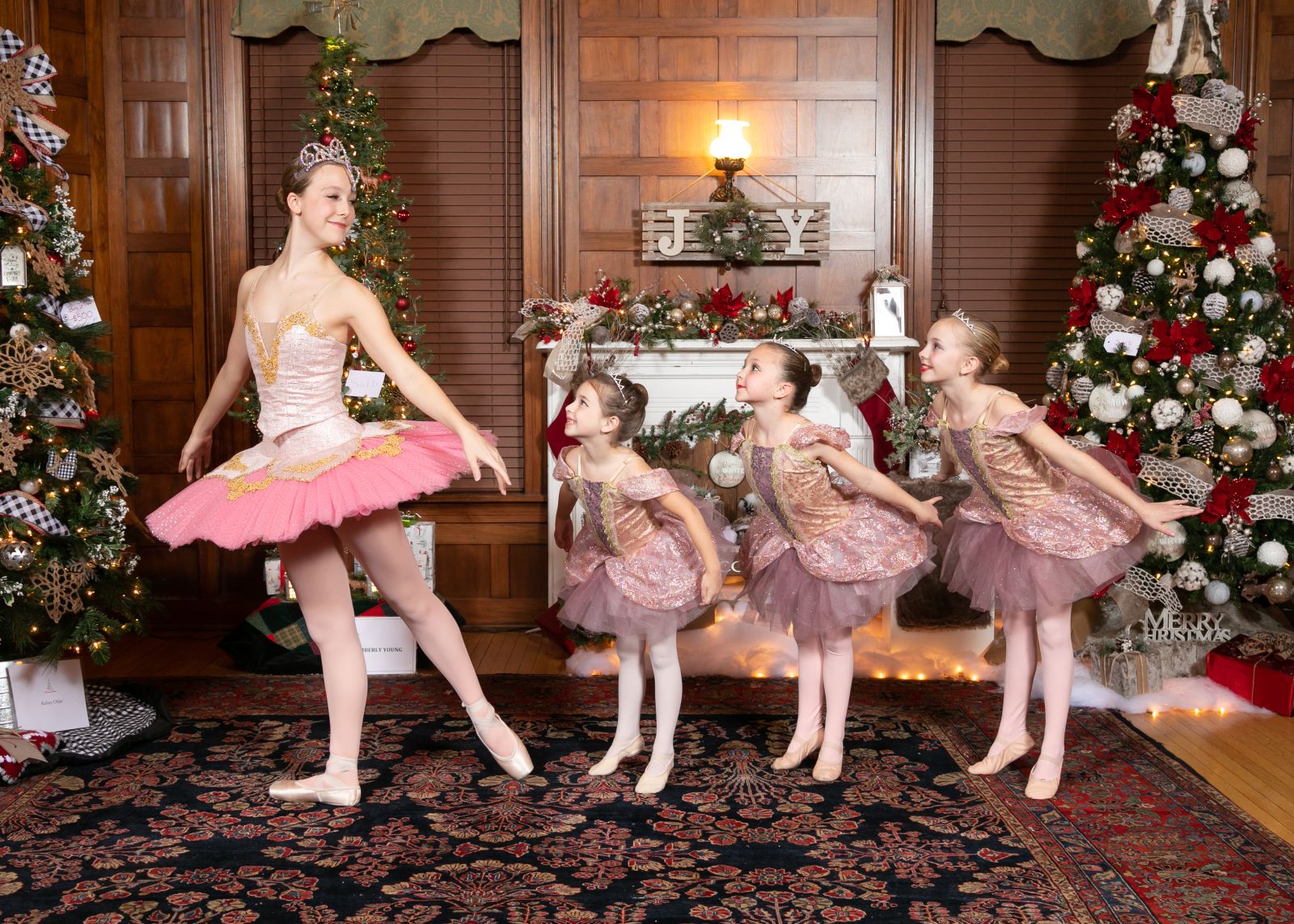 Three young girls in tutus lean toward a tall ballerina. They are in front of tw